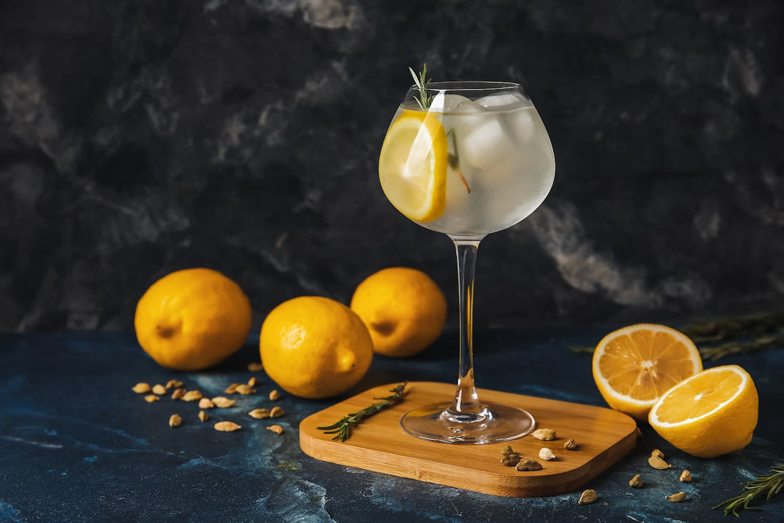 Copa de Tropical Gin decorada con limón y romero, rodeada de limones frescos y cardamomo, sobre una base de madera