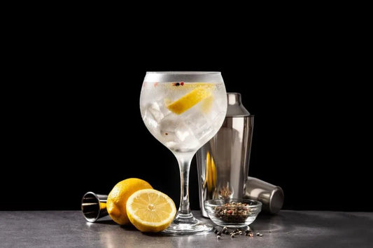 Copa de gin tonic servida con hielo, rodaja de limón y granos de pimienta, acompañada de limones frescos, botánicos en un recipiente pequeño y utensilios de coctelería sobre una superficie gris, con fondo negro