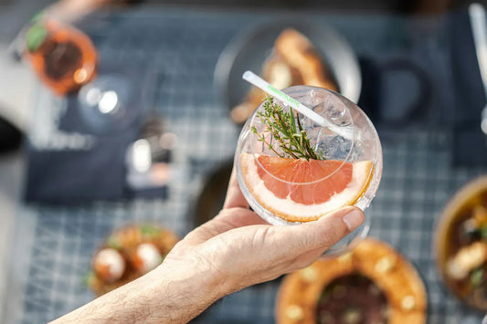Mano sosteniendo una copa de gin tonic adornada con una rodaja de pomelo rosado, ramitas de hierbas aromáticas y una pajilla reutilizable, con un fondo desenfocado que muestra una mesa con platos y bebidas