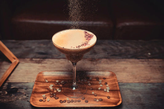Un cóctel de café servido en una copa elegante, decorado con cacao en polvo espolvoreado en el aire y granos de café sobre una bandeja de madera, con un fondo rústico de madera
