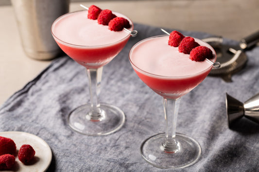 2 cócteles Clover Club servidos en una copa martini, decorados con frambuesas, sobre un mantel gris