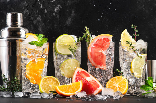 De izquierda a derecha, coctelera plateada, 4 vasos con hielo, cada vaso tiene una fruta distinta, naranja, limón sutil, pomelo, limón, sobre una mesa con más hielo y fruta, sobre un fondo negro