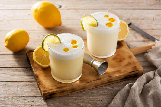 Mesa de madera, sobre esta se encuentran limones repartidos, una tabla de madera, sobre esta un jigger y dos cócteles pisco sour terminados.