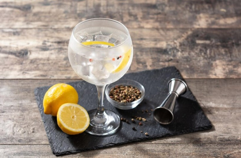 "Gin tonic en copa con hielo, limón, pimienta y especias sobre pizarra negra en mesa rústica