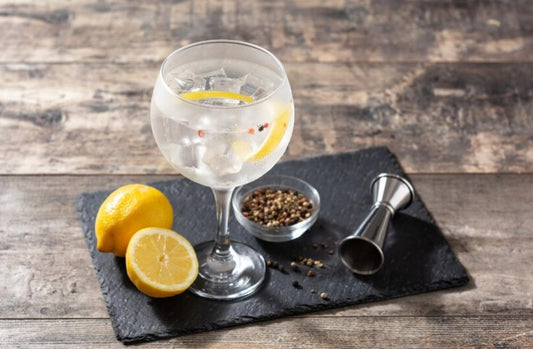 "Gin tonic en copa con hielo, limón, pimienta y especias sobre pizarra negra en mesa rústica