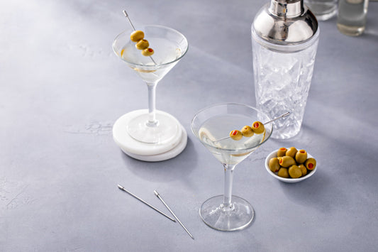 Dos copas de martini con aceitunas decorativas sobre una mesa gris, acompañadas de un mezclador metálico y un pequeño plato de aceitunas.