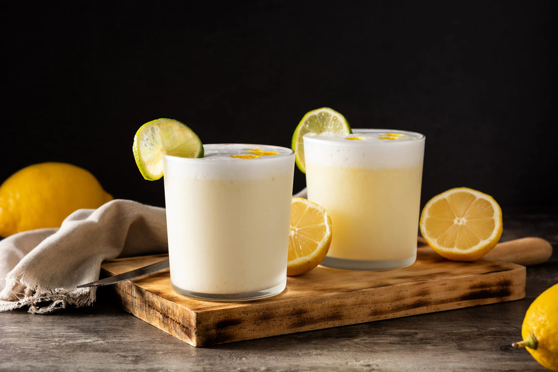 Dos cócteles de Pisco Sour con rodajas de limón, presentados en vasos sobre una tabla de madera, con limones frescos alrededor en un fondo oscuro